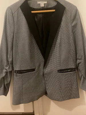 Rutig kavaj från H&M, storlek 42 - Snygg rutig kavaj från H&M i svart och vitt med markerade slag i svart tyg. Två fickor framtill med dragkedjor ger en cool touch. Perfekt för dig som vill ha en stilren men ändå edgy look. Passar till både jeans och kostymbyxor.