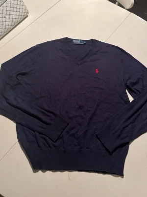 Mörkblå v-ringad tröja från Polo Ralph Lauren - Klassisk mörkblå v-ringad tröja från Polo Ralph Lauren i bomull. Tröjan har långa ärmar, ribbade muddar och den ikoniska röda loggan broderad på bröstet. Perfekt till jeans eller skjorta för en clean och stilren look.