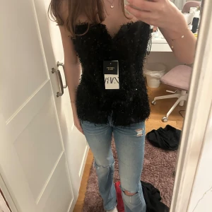 Svart paljettkorsett från Zara - Säljer en svart korsett från Zara med paljetter och fluffig textur. Toppen är axelbandslös och har knappar framtill. Perfekt för dig som vill sticka ut med en glittrig och trendig look. Sitter tight och formar kroppen snyggt.