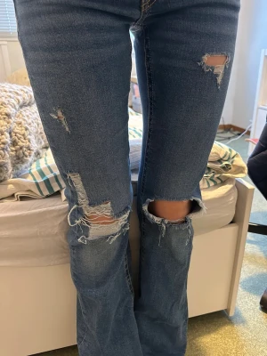 Blå bootcut jeans med hål på knäna - Säljer ett par blå jeans med bootcut passform och slitningar över knäna och låren. Jeansen har låg midja, klassiska fem fickor och dubbla knappar i linningen. Passar mig som är 168 perfekt. 