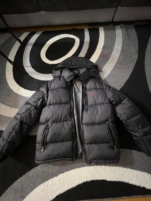 Svart pufferjacka från Polo Ralph Lauren - Säljer en svart pufferjacka från Polo Ralph Lauren i storlek M. Jackan har huva, två dragkedjeförsedda fickor och den klassiska röda loggan på bröstet. Perfekt för kalla dagar och riktigt skön att bära. Jag har aldrig använt den, jag fick den som julklapp!