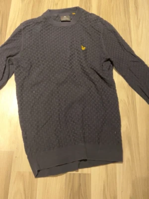Mörkblå tröja från Lyle & Scott - Säljer en mörkblå långärmad tröja från Lyle & Scott med diskret rutmönster och gul logga på bröstet. Tröjan har rund halsringning och är tillverkad i mjukt material som passar perfekt till höst och vinter.