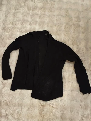 Svart stickad kofta från H&M - En svart, öppen stickad kofta från H&M i storlek M. Koftan har lång ärm och är tillverkad i ett mjukt, luftigt material som passar perfekt för lager på lager. Enkel och stilren design som funkar till det mesta.