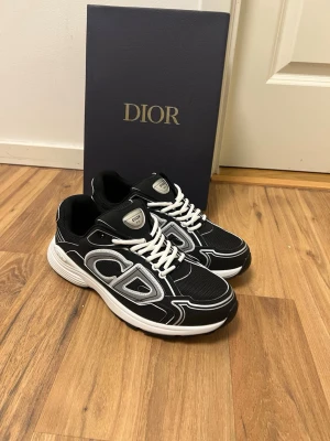 Dior b30 - Snygga svarta Dior sneakers med vita och grå detaljer samt tydlig CD-logga på sidan. Skorna har vit sula, svarta meshpaneler och vita skosnören. Perfekta för dig som vill ha en exklusiv och trendig look.