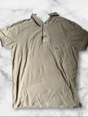 Beige Moncler pikétröja - Snygg beige pikétröja från Moncler med klassisk krage och knäppning framtill. Tröjan har Moncler-logga på bröstet och detaljer i vitt, rött och marinblått på krage och ärmslut. Tillverkad i mjuk bomull och har korta ärmar.