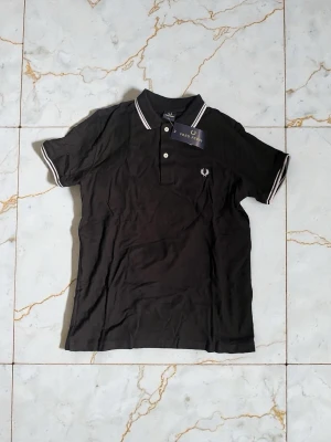 Svart piké från Fred Perry - Klassisk svart pikétröja från Fred Perry med vit logga på bröstet och vita ränder på krage och ärmslut. Tröjan har korta ärmar, två knappar framtill och är gjord i mjuk bomull. Perfekt till en clean och sportig stil.