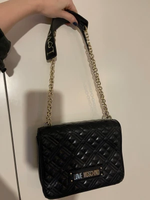 Svart quiltad axelväska Love Moschino - Säljer en svart quiltad axelväska från Love Moschino med guldfärgade kedjor och detaljer. Väskan har ett stort Love Moschino-emblem framtill och vadderad axelrem med logga. Tillverkad i skinnimitation och har en lyxig vibe.