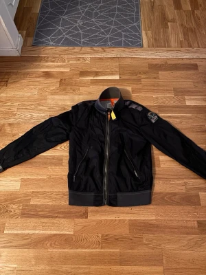 Svart bomberjacka från Parajumpers - Säljer en svart bomberjacka från Parajumpers i storlek S. Jackan har klassisk bomberstil med ribbade muddar, dragkedja framtill och två snedställda fickor med dragkedja. Detaljer som orange insida och ikonisk PJS-patch på ärmen. Perfekt för dig som vill ha en stilren och sportig look.