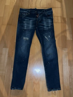Dsquared2 blå distressed jeans - Snygga blå jeans från Dsquared2 med slitna detaljer, färgstänk och trasiga partier på benen. Klassisk femficksmodell med röd Dsquared2-tag vid gylfen och patch bak. Jeansen har en smal passform och cool tvättad look.