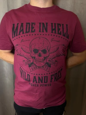 Vinröd t-shirt med dödskalletryck - Cool vinröd t-shirt med stort svart tryck framtill som visar en dödskalle med vingar och texten 'MADE IN HELL' och 'WILD AND FREE'. T-shirten har rund hals och korta ärmar, perfekt för dig som gillar edgy stil.