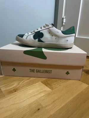 Vita och gröna sneakers The Gallerist - Snygga vita sneakers från The Gallerist med gröna detaljer och en stor grön klöver på sidan. Skorna har snörning, rund tå och platt sula. Ovandelen är i skinn och tån samt hälen har gröna partier i mocka. Perfekta för dig som vill sticka ut med en unik design.