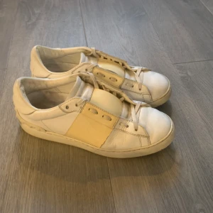 Valentino Rockstud sneakers i vitt och beige - Snygga Valentino Rockstud sneakers i vitt skinn med beige lackad panel över snörningen. Platt sula med ikoniska nitar baktill och rund tå. Skorna har snörning och en stilren design som sticker ut med sina detaljer.