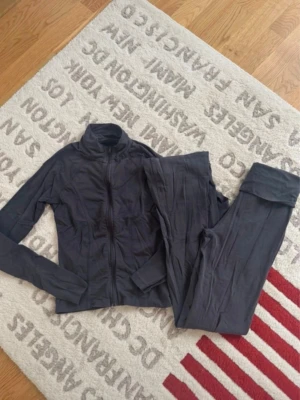 Mörkgrå kofta och byxor från Gina Tricot - Säljer en mörkgrå kofta och byxor från Gina Tricot med dragkedja framtill och hög krage. Dem är i mjukt bomullsmaterial och har en enkel, stilren design utan mönster eller extra detaljer. 