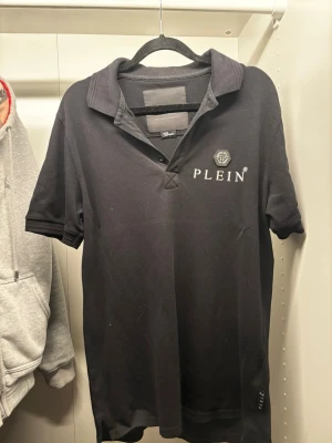 Svart Philipp Plein pikétröja - Säljer en svart pikétröja från Philipp Plein med klassisk krage och korta ärmar. Tröjan har broderad logga och emblem på bröstet samt diskret etikett bak i nacken. Tillverkad i bomull för en skön och stilren look.