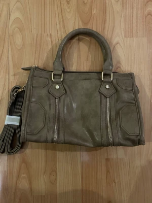 Brun/khaki handväska med gulddetaljer - Snygg handväska med guldfärgade detaljer och dragkedjor framtill. Väskan har både handtag och en lång axelrem som går att ta av. Mått finns på bilderna och passar perfekt till alla dina essentials. Pris går att diskutera💞