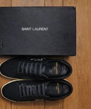 Svarta Saint Laurent sneakers - Svarta sneakers från Saint Laurent med vit handskriven logga på sidan och klassisk snörning. Skorna har en beige platt sula och ovandel i textil med detaljer i skinn grova svarta skosnören. Perfekta för dig som centered streetstyle och vill ha något unikt.