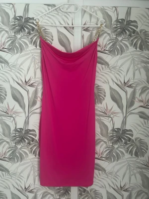 Rosa kortklänning med guldkedjor - Supersnygg rosa kortklänning från By Swan med tunna axelband i guldfärgad kedja. Klänningen har en draperad urringning och är figurnära i modellen. Perfekt för dig som vill sticka ut och älskar färg! Materialet känns mjukt och följsamt.