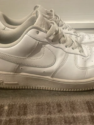 Vita Nike Air Force 1 sneakers - Klassiska helvita Nike Air Force 1 sneakers med låg profil och snörning. Skorna är tillverkade i skinn med perforerade detaljer på tån och har den ikoniska swoosh-loggan på sidan. Perfekta för en clean och stilren look.