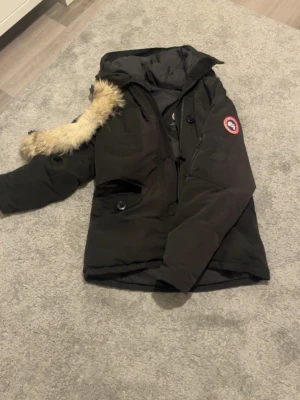  Äkta Svart Canada Goose med päls - Svart parkajacka från Canada Goose med ikonisk röd logga på ärmen och huva med beige pälsdetalj. Jackan har dragkedja, knappar och flera fickor. Perfekt för kalla dagar med sin tjocka dunfyllning och snygga design. OBS, jackan är äkta.