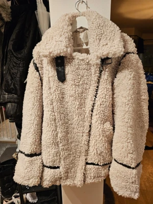 Beige teddyjacka med svarta detaljer - Säljer en fluffig beige teddyjacka med stora slag och svarta detaljer längs dragkedja och sömmar. Jackan har en oversized look och stängs med sned dragkedja. Perfekt för kalla dagar när du vill vara både varm och snygg.