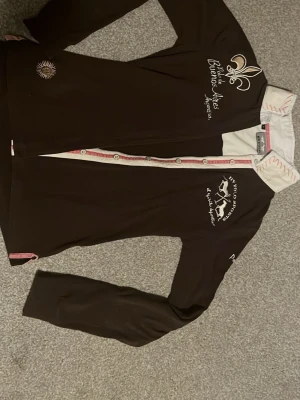 Brun långärmad ridtröja från HV Polo - Säljer en brun långärmad ridtröja från HV Polo med vit krage och broderade detaljer framtill. Skjortan har knappar hela vägen. Perfekt för dig som gillar ridning!