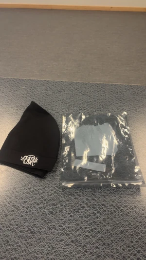 Svart mössa från Syna world  - Säljer en svart mössa från syna world med vit broderad logga framtill. Mössan är enkel och stilren med mjukt material som sitter skönt på huvudet. Perfekt för kalla dagar och passar till de flesta outfits.