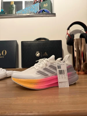 Adidas Ultraboost med färgad sula - Säljer ett par Adidas Ultraboost sneakers i vitt med mesh-överdel och klassiska tre ränder i grått. Sulan har en snygg gradient i orange, rosa och gul som verkligen poppar. Perfekta för dig som vill ha både stil och komfort till träningen.