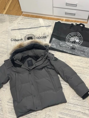 Grå Canada Goose Wyndham - Säljer en stilren grå Canada Goose vinterjacka i mycket fint skick. Har varm dunfyllning, huva med pälskant och klassisk Canada Goose-logga på ärmen. Perfekt för kalla dagar och för dig som vill ha en både snygg och riktigt varm jacka