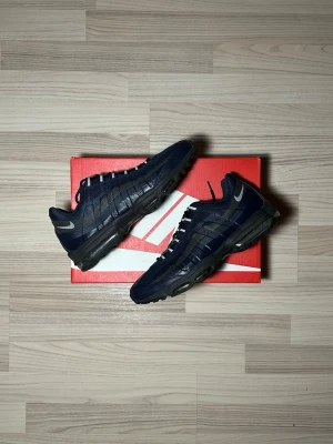 Nike Air Max 95 marinblå sneakers - Säljer ett par Nike Air Max 95 sneakers i marinblått med svarta och grå detaljer. Skorna har synlig Air-dämpning i sulan, klassisk snörning och mesh- samt syntetpaneler för en sportig look. Swoosh-logga på sidan och vågiga linjer längs ovandelen. Köpbevis finns (Zalando)