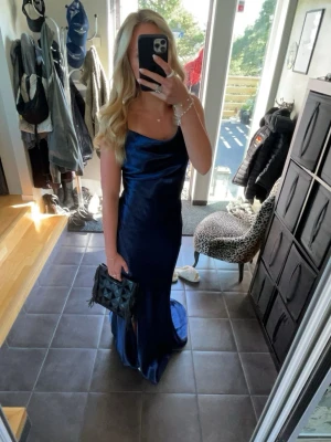 Mörkblå långklänning i satin - Säljer en elegant mörkblå långklänning i satin med smala axelband och öppen rygg. Klänningen har en snygg draperad urringning och figurnära passform som ger en lyxig känsla. Perfekt för bal eller festliga tillfällen! Storleken är 36/S men passar också M/38. Endast använd en gång på balen i 9:an!