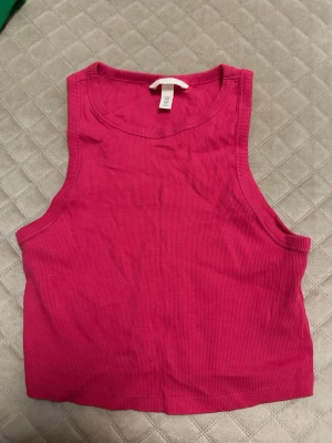 Ribbad rosa croppad linnetopp H&M - Säljer en ribbad croppad linnetopp från H&M i en stark rosa färg. Toppen är ärmlös med breda axelband och har en enkel, sportig design. Perfekt till sommaren och lätt att matcha med jeans eller kjol.