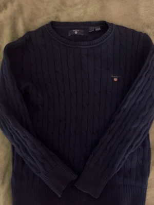 Mörkblå kabelstickad tröja från GANT - Kabelstickad mörkblå tröja från GANT med rund hals och diskret logga broderad på bröstet. Tröjan har långa ärmar och ribbade muddar vid ärmslut och nederkant. Perfekt för dig som gillar klassisk och stilren look. Knappt använd