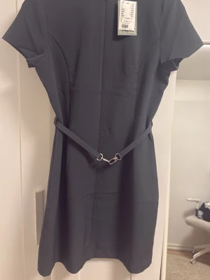 Grön kort klänning med bälte - Snygg grön kort klänning med korta ärmar och ett avtagbart bälte i midjan. Klänningen har en enkel, stilren design och är tillverkad i ett mjukt material som ger en clean look. Perfekt för dig som gillar minimalistisk stil.