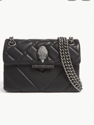 Svart quiltad axelväska med kedja - Snygg svart axelväska från Kurt Geiger i quiltat skinn med silverfärgad kedja och ikoniskt örnhuvud som spänne. Väskan har en kompakt form och coola detaljer som ger en lyxig känsla. Perfekt för dig som vill ha en statementväska med edge.