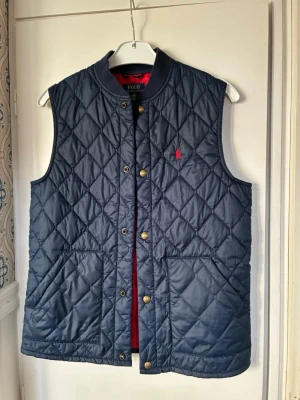 Mörkblå quiltad dunväst från Polo Ralph Lauren - Snygg mörkblå quiltad dunväst från Polo Ralph Lauren med klassisk röd logga på bröstet. Västen har tryckknappar i guld, två stora fickor framtill och en hög krage. Perfekt att slänga över en hoodie eller tröja för extra värme.
