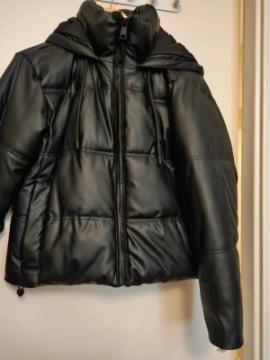 Svart pufferjacka från Zara  - Säljer en svart pufferjacka från Zara i snyggt syntetmaterial med skinnlook. Jackan har huva, dragsko nertill och dragkedja framtill. Perfekt för kalla dagar och ger en cool vibe till din outfit.