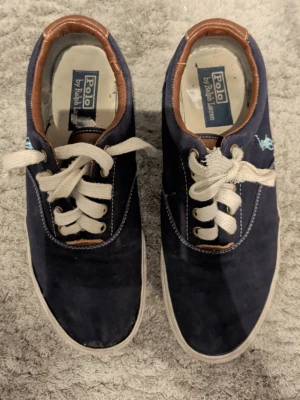 Marinblå sneakers från Polo Ralph Lauren - Snygga marinblå sneakers från Polo Ralph Lauren med vita snören och bruna skinn-detaljer vid häl och öppning. Skorna har broderad logga i ljusblått på sidan och klassisk rund tå. Ovandelen är i slitstark bomull och sulan är vit.