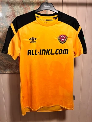 Dynamo Dresden Umbro 2021/22 home shirt, size M - Mycket bra skick.  #germany #bundesliga #bundesliga2 #umbro  Dynamo Dresden fotbollströja med tryck av 'ALL-INKL.COM' på framsidan och årtalet '1953' på baksidan. Tröjan har en guldfärgad bas med svarta detaljer på axlarna och kragen. Klubbmärket och Umbro-logotypen är placerade på bröstet.