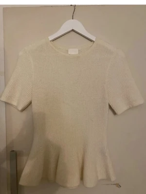 Beige ribbad stickad topp - Säljer en beige ribbad stickad topp med korta ärmar och rund hals. Toppen har en lätt utsvängd nederkant som ger en snygg siluett. Perfekt att styla med jeans eller kjol för en clean look.