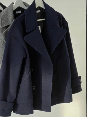 Mörkblå kappa med knappar - Säljer en mörkblå skepparkavaj med bred krage och dubbelknäppning framtill. Jackan har stora fickor, uppvikta ärmslut med knappdetalj och en rak, loose passform. Materialet är tjockt och känns som ull, perfekt för kyligare dagar. Storlek xs/s