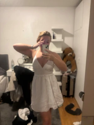 Vit kort sommarklänning med smala band - Supersöt vit kort klänning med smala axelband och v-ringning. Klänningen har en lätt rynkad kjol och öppen rygg med resårdetalj. Perfekt för varma dagar och har en luftig känsla. Materialet känns som bomull och den har en diskret randig struktur.