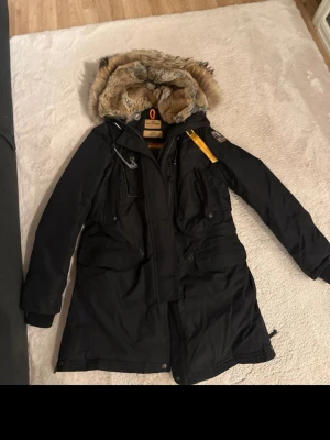 Svart Parajumpers parkas med päls - Säljer en svart Parajumpers parkas med stor huva fodrad i mjuk pälsimitation och snygga detaljer som gul rem och patch på ärmen. Jackan har flera fickor, dragkedja och knappar framtill. Perfekt för kalla dagar och riktigt varm.