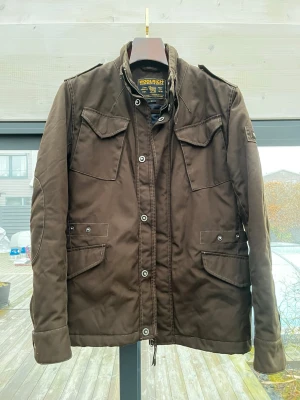 Woolrich Jacka - Säljer en brun fieldjacka från Woolrich med fyra stora fickor framtill, axelklaffar och quiltat innerfoder. Jackan har både dragkedja och knappar framtill för extra skydd mot vind. Perfekt för dig som vill ha en stilren och funktionell jacka.