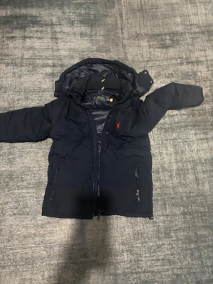 Mörkblå dunjacka från Polo Ralph Lauren - Säljer en mörkblå dunjacka från Polo Ralph Lauren med huva och dragkedja framtill. Jackan har två fickor med dragkedja och klassisk röd logga på bröstet. Perfekt för kalla dagar och riktigt snygg till streetwear-stilen. Köpte den när jag var i USA och använde aldrig den.