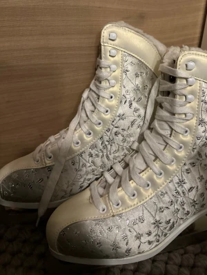 Tecnopro Vita skridskor med blommigt mönster - Unika vita skridskor med silverfärgat blommigt broderi och snörning. Utsidan är i glansigt satinliknande material och insidan är fodrad med mjuk fuskpäls.  Endast använt några enstaka gånger.