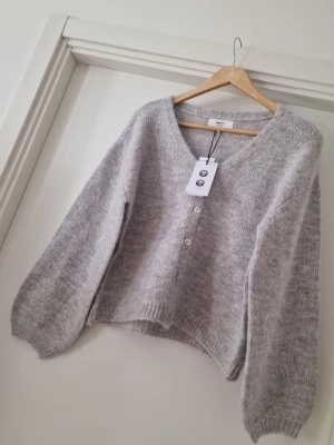 Ljusgrå kofta från Gina Tricot - Supermjuk ljusgrå kofta från Gina Tricot i ullblandning. Koftan har v-ringning, tre knappar framtill och ballongärmar för en chill vibe. Perfekt att slänga över en topp när det är kyligt ute. Storlek XS. Ny med prislapp