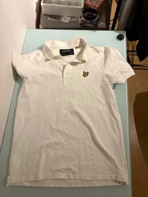 Vit pikétröja från Lyle & Scott S - Vit pikétröja från Lyle & Scott i storlek S. Klassisk design med krage, korta ärmar och knappar framtill. Tröjan har den ikoniska gula fågel-loggan broderad på bröstet. Tillverkad i bomull för en skön och luftig känsla.