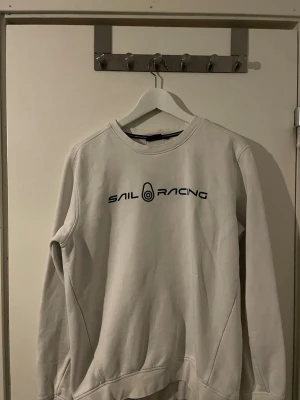 Vit Sail Racing sweatshirt i bomull - Säljer en stilren vit sweatshirt från Sail Racing i storlek M. Tröjan har rund halsringning, svart logotyptryck på bröstet och är tillverkad i mjuk bomull. Perfekt för dig som gillar en clean och sportig look.