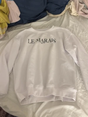 Vit sweatshirt Le Marais - Vit sweatshirt med svart text 'LE MARAIS' på bröstet. Tröjan har rund halsringning och ribbade muddar vid ärmslut och nederkant. Perfekt för en chill och stilren look. Mjuk insida för extra komfort.