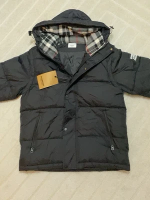 Svart dunjacka från Burberry - Säljer en svart dunjacka från Burberry med klassiskt rutigt foder i huvan. Jackan har dragkedja, tryckknappar och två fickor med dragkedja framtill. Perfekt för kalla dagar och har en modern, puffig look. Helt ny kontakta vid frågor 